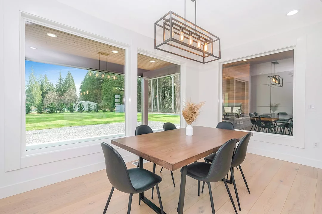 A modern dining room with a wooden table, six black chairs, large windows overlooking a green yard, and a geometric light fixture overhead offers a touch of luxury to your everyday lifestyle.