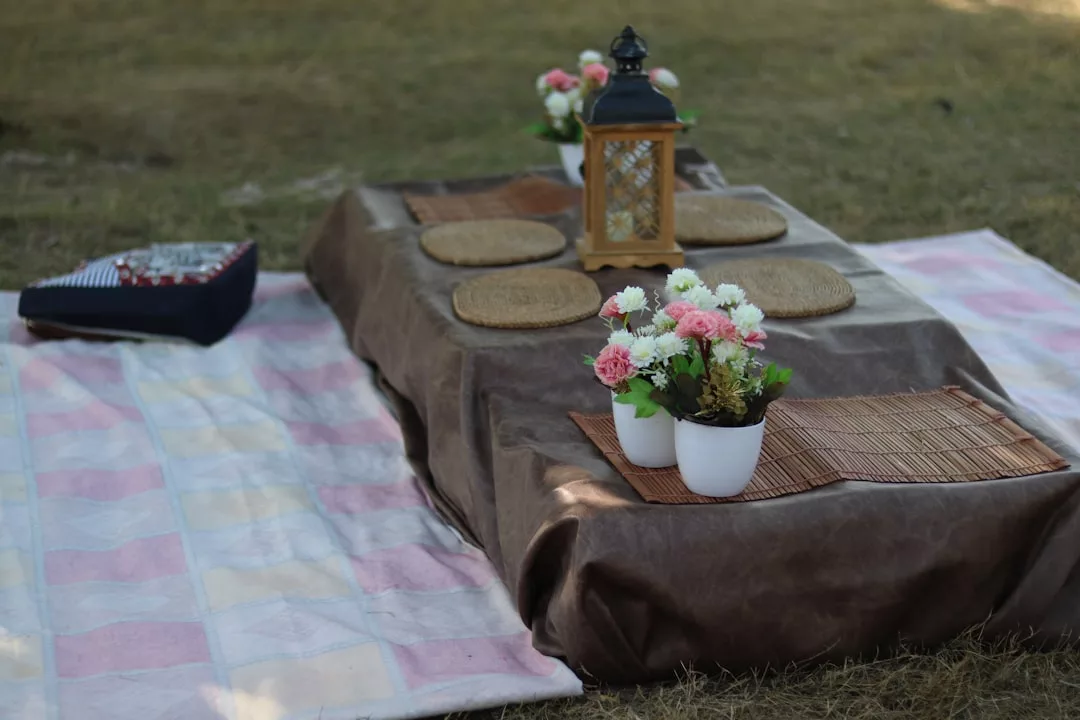 A low table with a brown cloth, wicker placemats, a lantern, flower pots, and a pink-checkered blanket creates a luxury picnic setup on grass—perfect for enjoying the relaxed Majorca lifestyle outdoors.