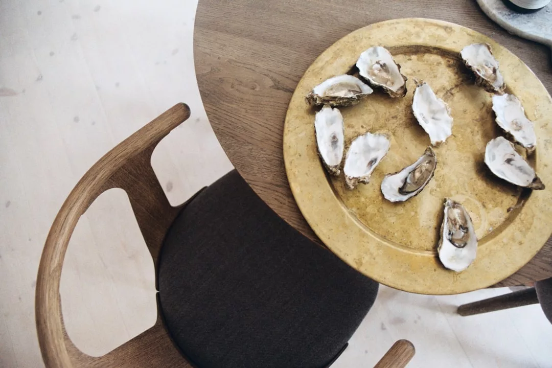 A round wooden table exudes luxury with a gold tray holding seven opened oyster shells, evoking a refined lifestyle next to a wooden chair with a dark cushion.