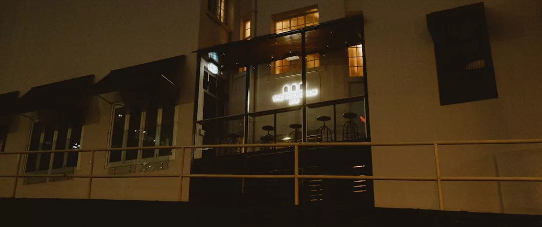 A glass-enclosed outdoor seating area with empty chairs and tables attached to a building at night, illuminated by indoor lights, evokes a sense of relaxed luxury and sophisticated lifestyle reminiscent of evenings in Majorca.