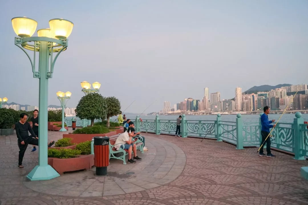People are fishing and relaxing on benches along a waterfront promenade, embracing a relaxed lifestyle with city buildings and mountains visible across the water in the background.
