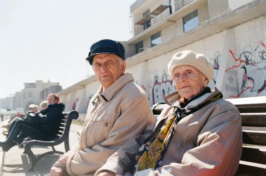 Two elderly people sit on a bench outdoors in Majorca, wearing light-colored coats and hats, with a graffiti-covered wall and other people in the background, capturing a unique blend of local lifestyle.