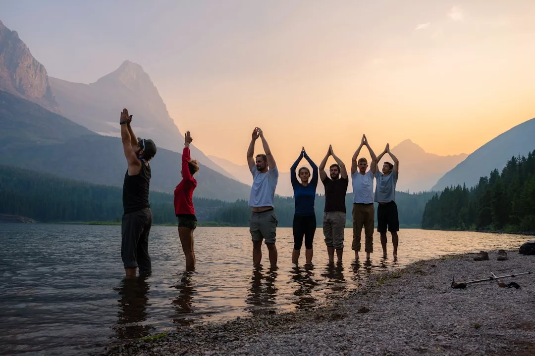Seven people stand in shallow water by a mountain lake at sunset, posing with arms raised in various positions, embracing a lifestyle of adventure, with mountains and trees in the background.