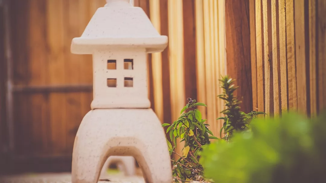 A small stone lantern stands next to green plants along a wooden fence, adding a touch of luxury to the serene garden—perfect for enjoying a tranquil Majorca-inspired lifestyle.