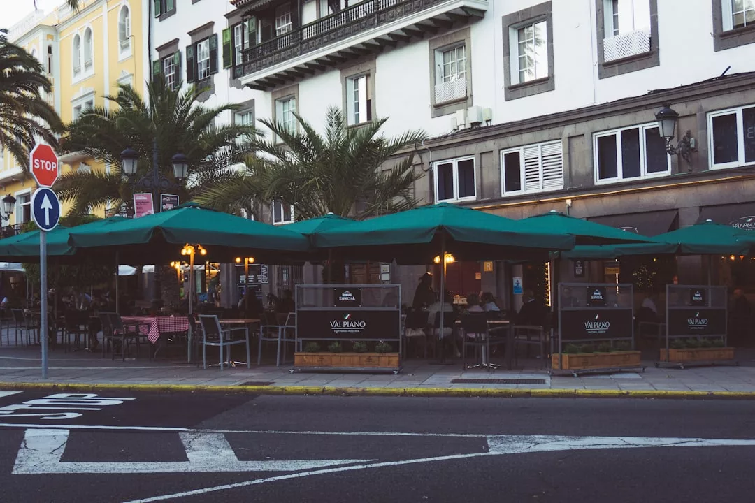Outdoor restaurant seating with green umbrellas lines a city street in Majorca, with palm trees and a multi-story building in the background, creating a scene that reflects luxury and a vibrant Mediterranean lifestyle.