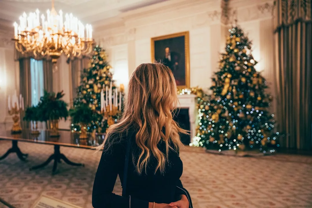 A woman with long blonde hair stands in an elegantly decorated room, surrounded by Christmas trees, chandeliers, and a large portrait on the wall, embodying a lifestyle of luxury.