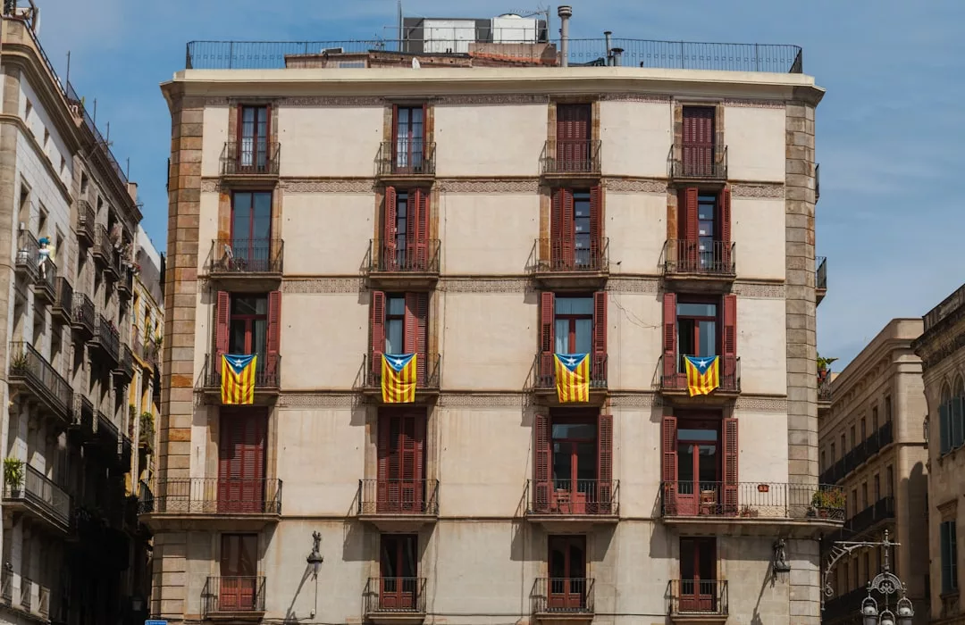 A beige four-story building with red shutters in Majorca displays five yellow, blue, and red flags hanging from balconies, hinting at a vibrant lifestyle. Neighboring buildings are partially visible.