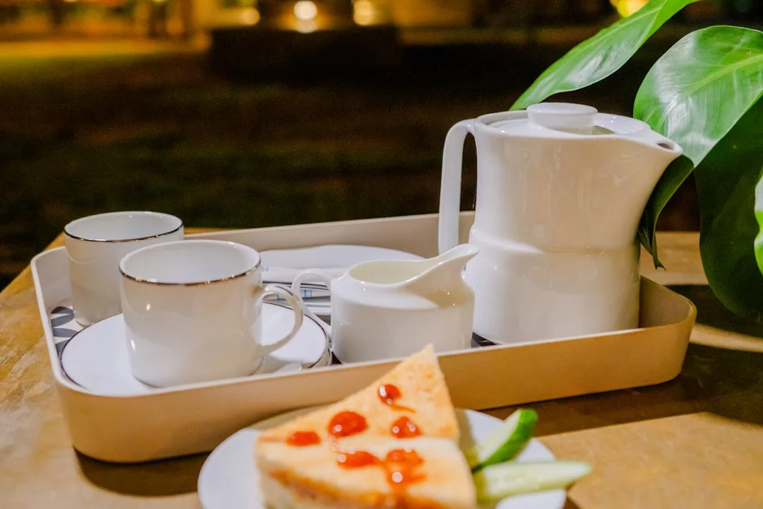 A white tray on a table holds a teapot, two cups and saucers, a small creamer, and utensils. In the foreground, a plate has a slice of cake with sauce and cucumber slices—an inviting touch of luxury, reminiscent of Majorca’s relaxed lifestyle.