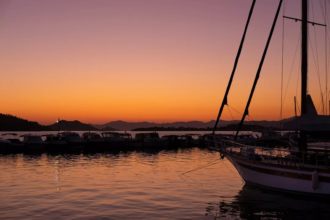 A sailboat and several docked boats are silhouetted against an orange and purple sunset over calm water with distant mountains, capturing the luxury lifestyle of Majorca.