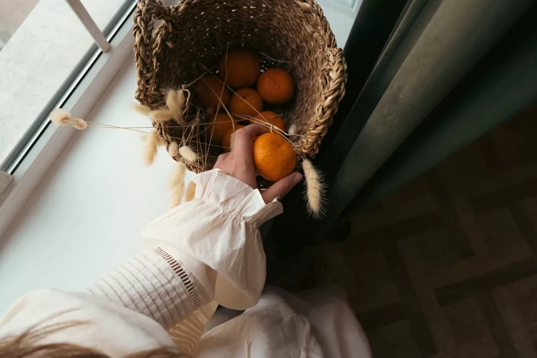 A person in a white blouse reaches into a woven basket filled with oranges and dried grasses near a window, capturing the relaxed lifestyle of Majorca.
