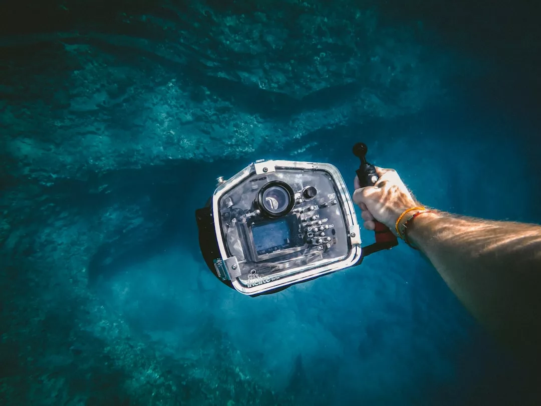 A person holds an underwater camera housing, pointing it towards the blue ocean floor off Majorca, their arm extended into the frame, capturing a glimpse of a luxurious lifestyle beneath the waves.