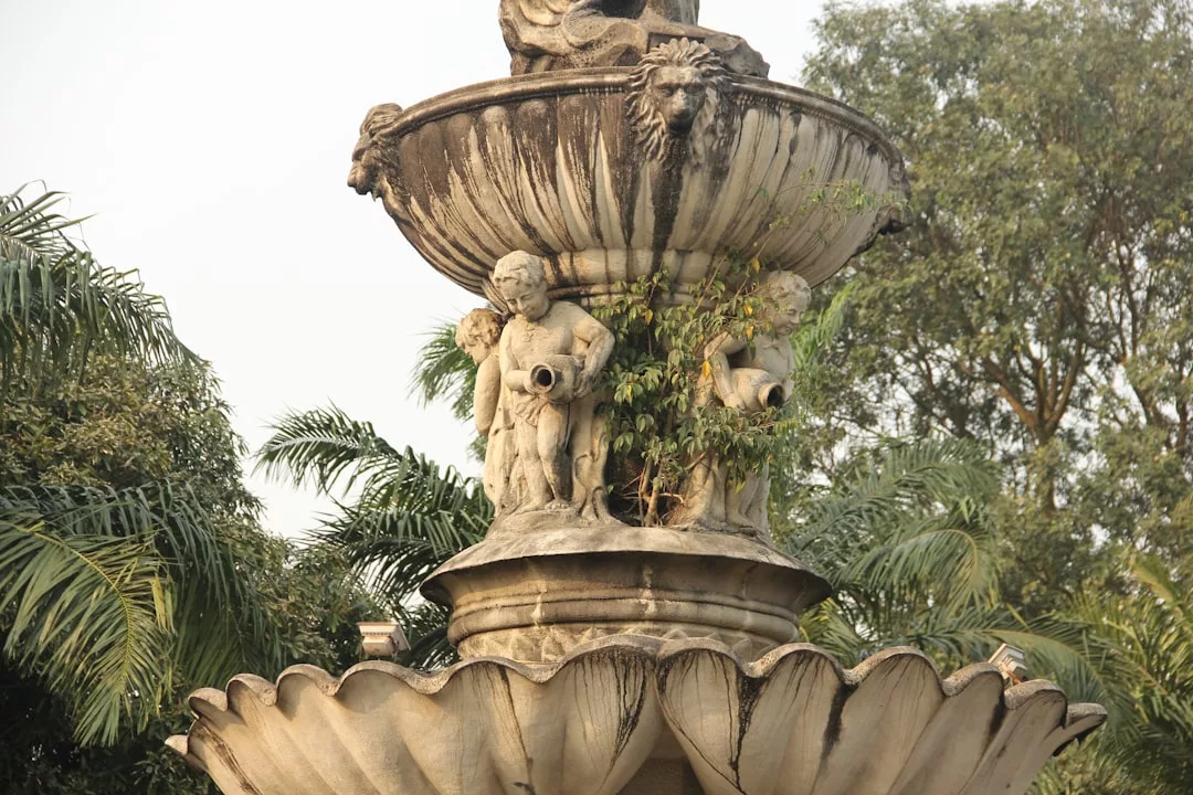 A stone fountain features cherub statues and lion heads, with plants growing from its upper tier, adding a touch of luxury to the lifestyle ambiance, surrounded by trees and palm fronds reminiscent of Majorca.