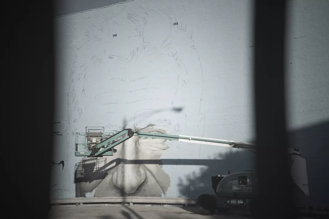A mural artist on a lift paints the lower part of a large face on a building wall in Majorca, with shadows and construction barriers in the foreground, adding vibrant flair to the island’s luxury lifestyle.