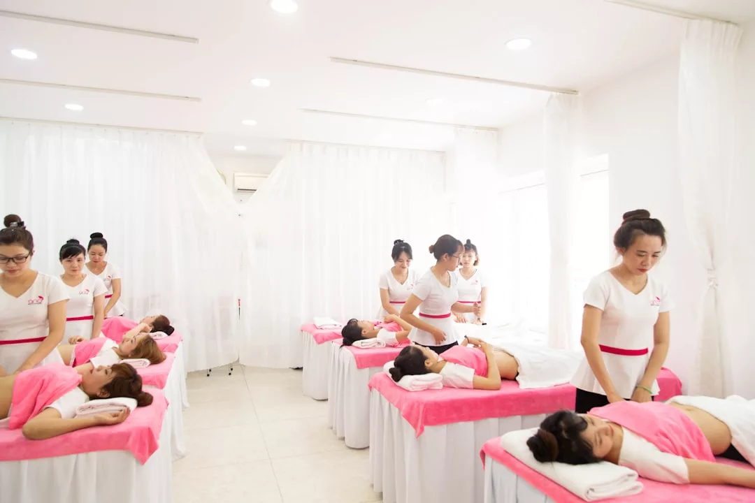 Several women enjoy luxury massages on pink-draped tables in a bright, white-curtained spa in Majorca, attended by staff in crisp white uniforms.