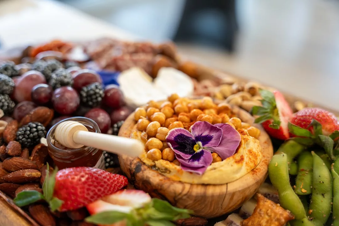 Experience a taste of luxury lifestyle with a wooden platter featuring assorted fruits, nuts, a bowl of hummus topped with chickpeas and an edible flower, honey dipper, sliced cheese, and fresh strawberries inspired by Majorca.