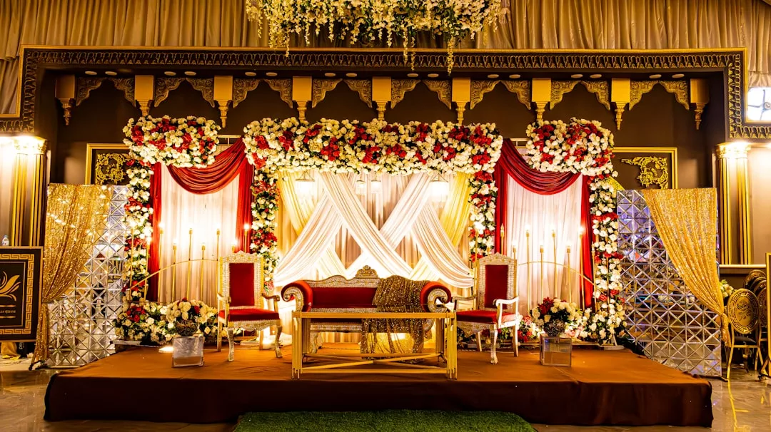 Ornate wedding stage decorated with red and white flowers, gold accents, draped fabric, and elegant chairs under warm lighting in a grand hall for a touch of luxury lifestyle.