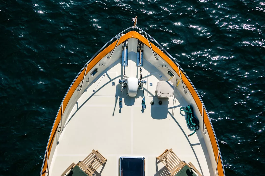 Aerial view of the bow of a luxury boat with wooden trim, deck chairs, coiled hose, and anchor equipment drifting on the blue waters of Majorca.