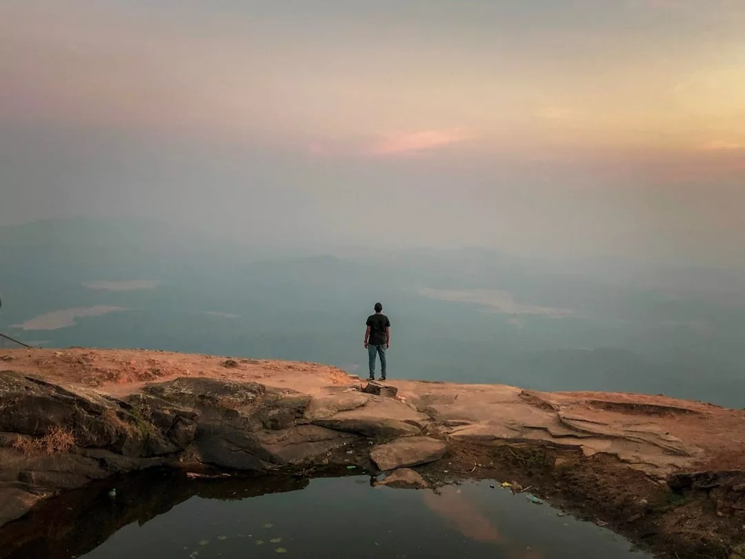 A person stands alone on a rocky cliff edge in Majorca, overlooking a hazy landscape of mountains, a river, and a cloudy sky at sunset—capturing the essence of serene luxury.