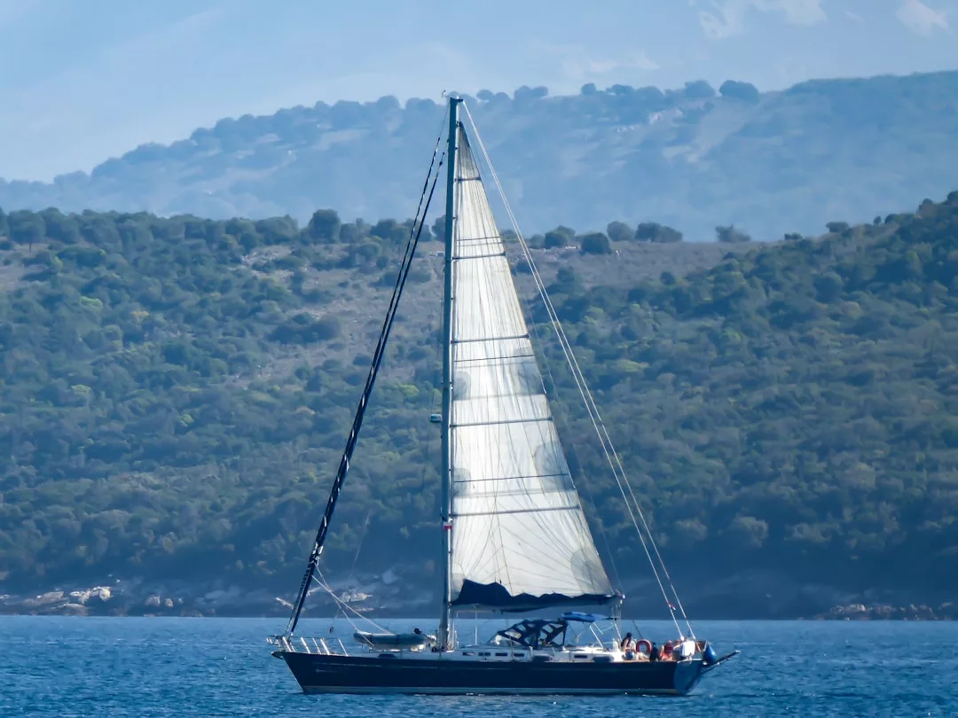 A luxury sailboat with its sails up floats on calm blue water near Majorca, with a green, hilly shoreline and mountains in the background, capturing the essence of an idyllic lifestyle.