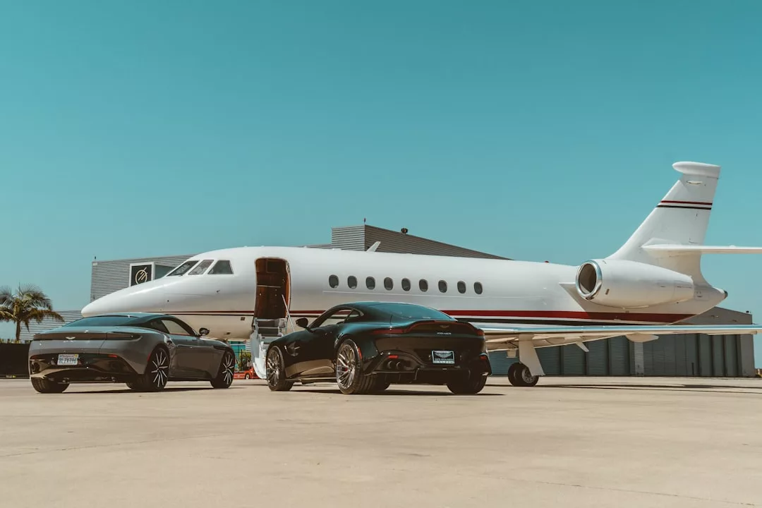 A private jet is parked on an airport tarmac in Majorca with its door open, beside two black luxury cars, in front of a hangar under a clear blue sky—a true statement of luxury lifestyle.