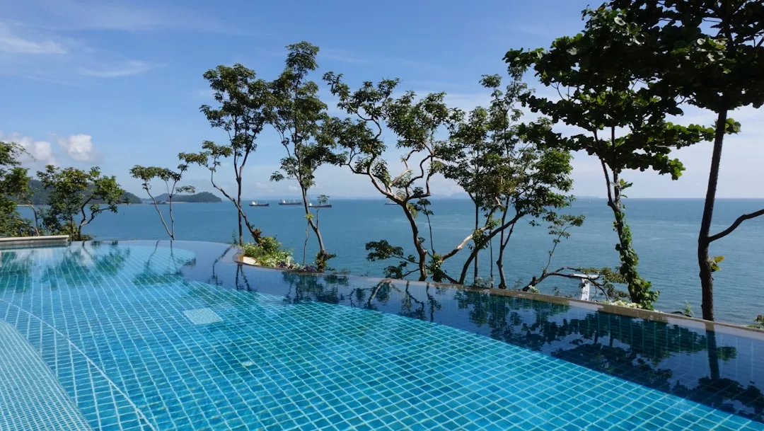 Infinity pool with blue tiles overlooking the ocean in Majorca, bordered by trees, with ships on the horizon under a clear, sunny sky—perfect for enjoying a luxury lifestyle.