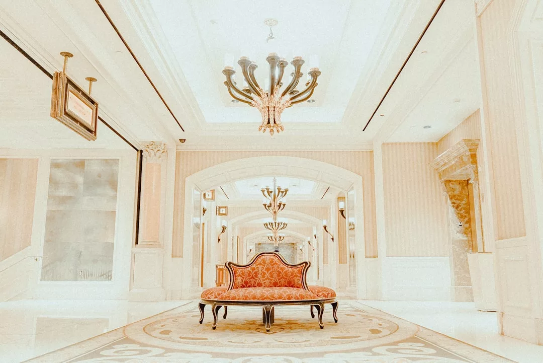 A luxurious hallway inspired by the Majorca lifestyle, featuring ornate chandeliers, a red upholstered bench on a patterned rug, and cream-colored walls and columns.