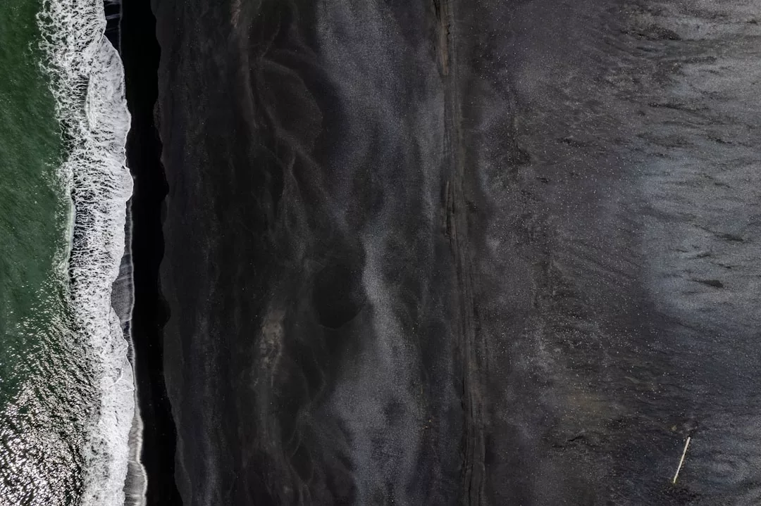 Aerial view of ocean waves meeting a dark sandy beach in Majorca, with green water on the left and black sand stretching across the image—capturing hints of luxury and relaxed lifestyle.