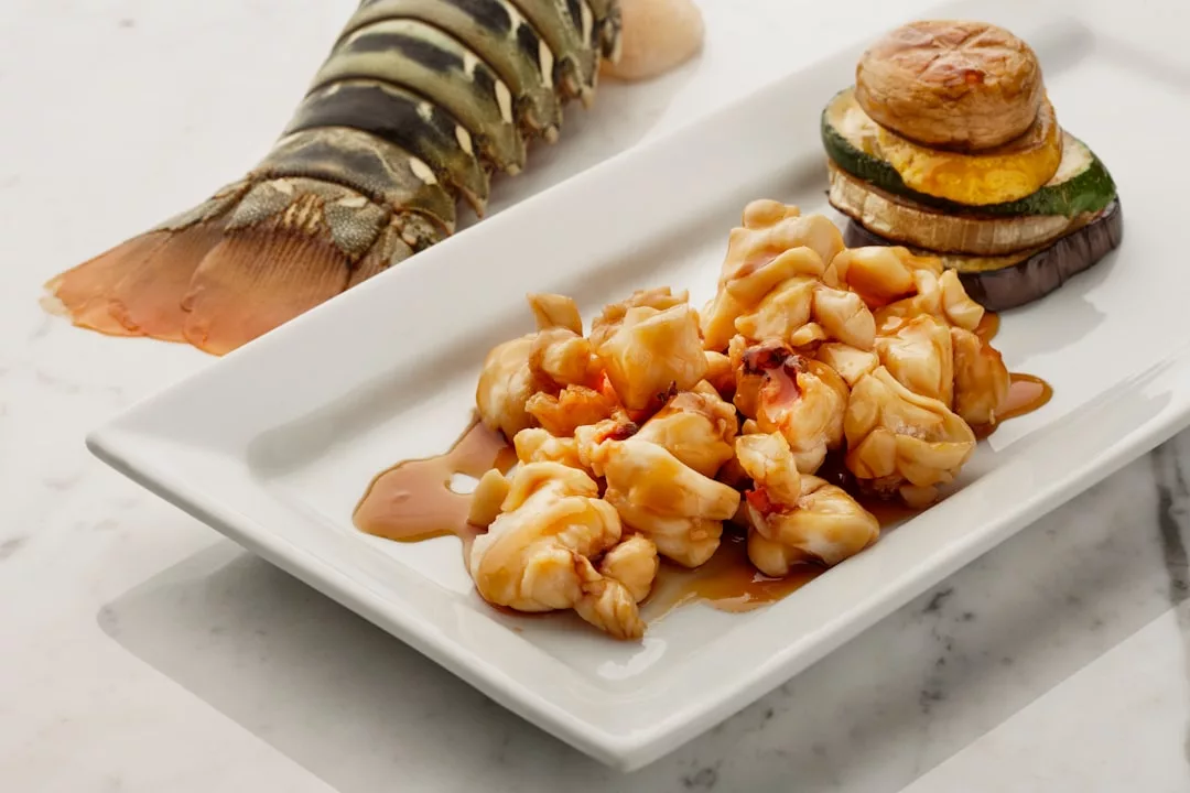 Experience luxury dining with a plate of cooked lobster tail meat and sauce, accompanied by grilled eggplant, zucchini, and a potato stack, garnished with a lobster shell—perfect for a refined lifestyle inspired by Majorca.