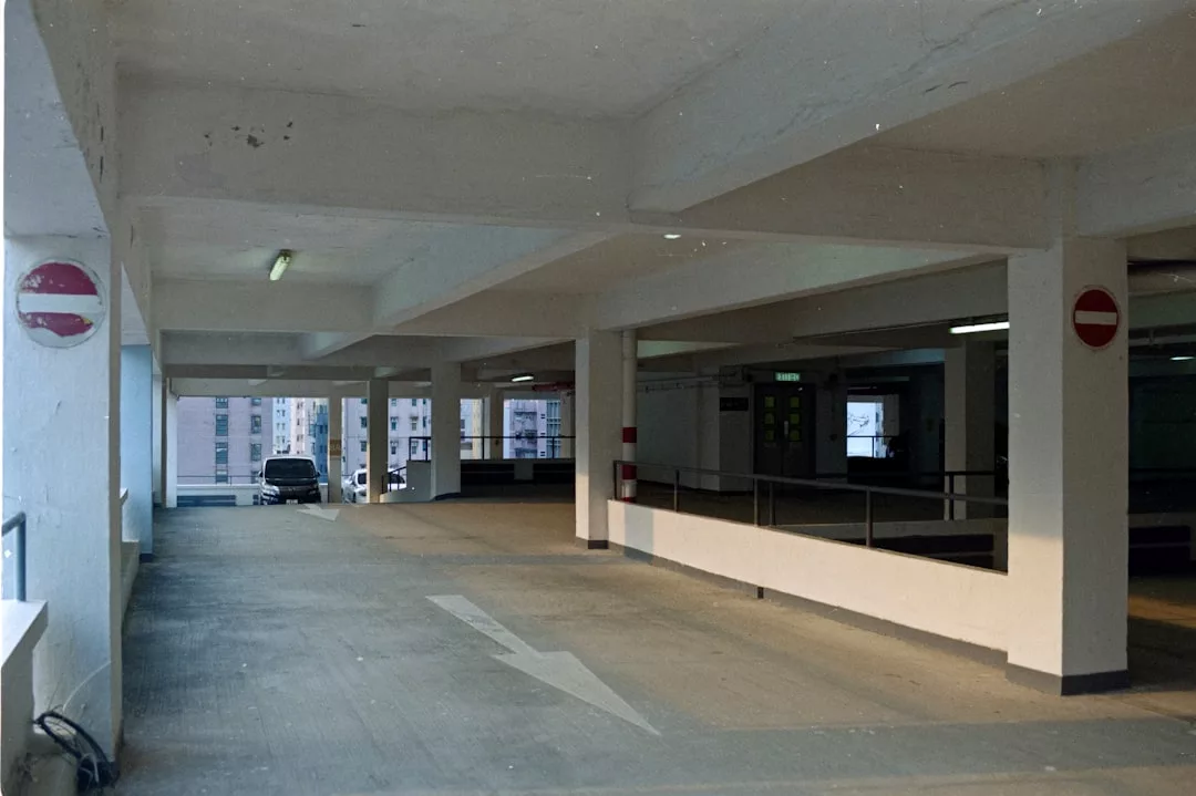Empty multi-story parking garage with white walls, support columns, overhead lighting, and no entry signs. A single luxury car is parked near the open side facing a building, adding a touch of exclusive lifestyle to the scene.