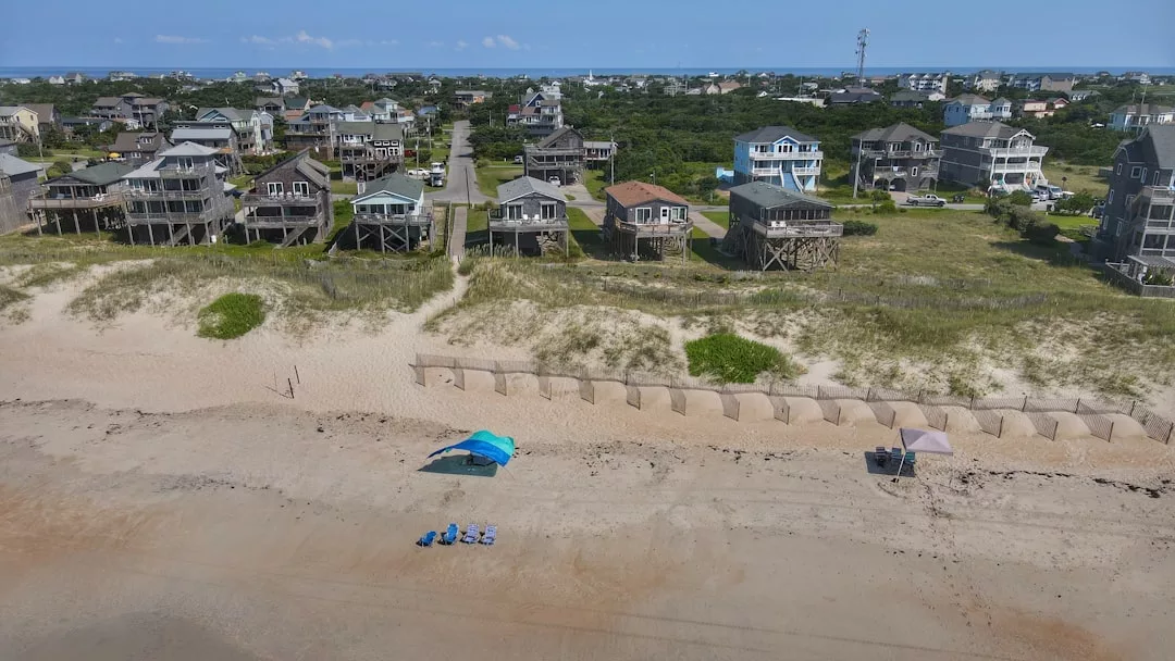 Aerial view of a sandy beach with a blue canopy and chairs, small dunes, and luxury houses on stilts lining the background with greenery and roads—perfect for enjoying a chic lifestyle reminiscent of Majorca.