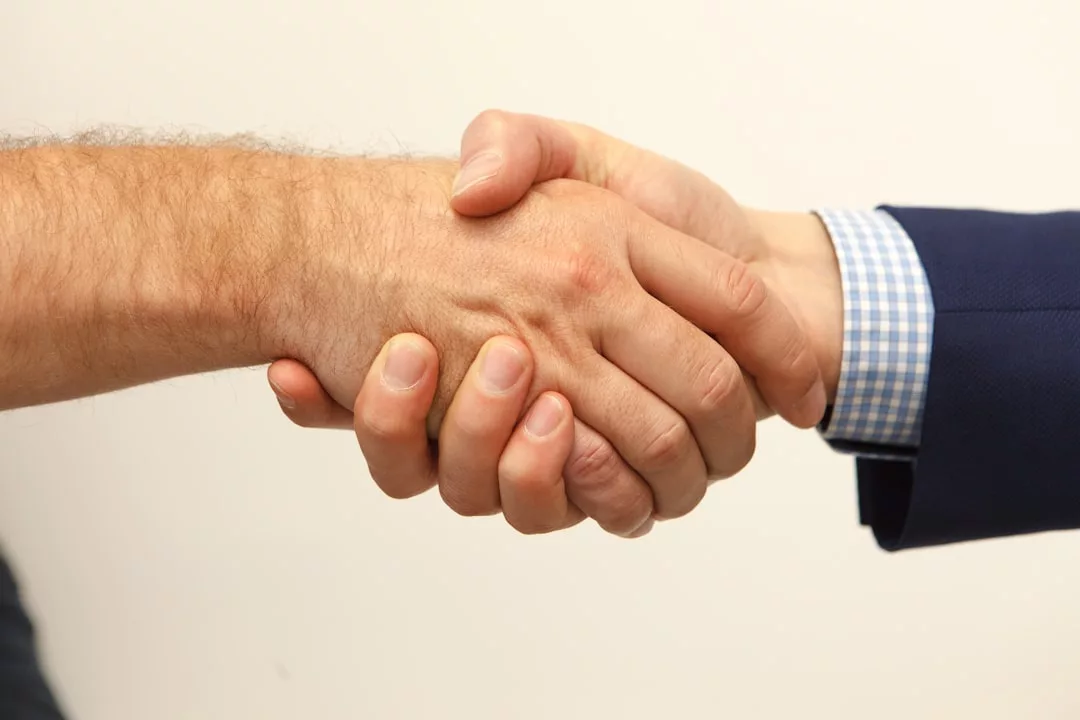 Two people shaking hands, one in a blue suit and checkered shirt, the other with a bare arm, set against a plain light background—capturing a moment of luxury lifestyle reminiscent of Majorca’s sophisticated charm.