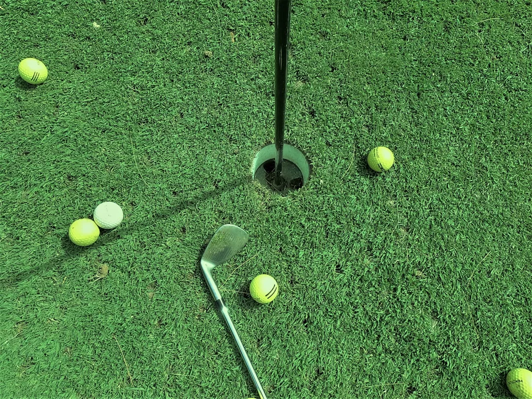A golf club and several yellow golf balls are scattered on luxury artificial green turf near a hole with a flagstick, capturing a stylish sporting lifestyle.