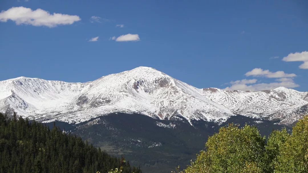 Snow-capped mountain under a clear blue sky with patches of green forest in the foreground, capturing the essence of luxury lifestyle inspired by Majorca’s breathtaking landscapes.