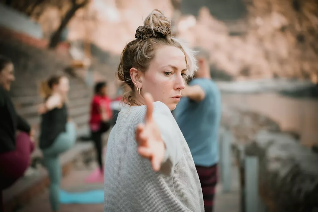 A woman practices a yoga pose outdoors in Majorca, extending her arm forward with a focused expression, while others join in the background, embracing a serene and luxurious lifestyle.