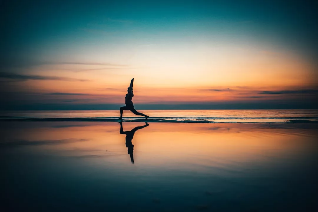 A person practices a yoga pose on a beach in Majorca at sunset, their silhouette and reflection visible against the colorful sky and water, capturing a serene moment of luxury lifestyle.