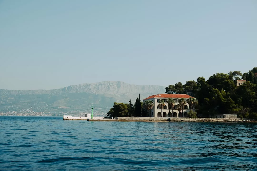 A luxury house with arched windows and a red roof sits by the water's edge in Majorca, surrounded by trees, with mountains visible in the hazy background.