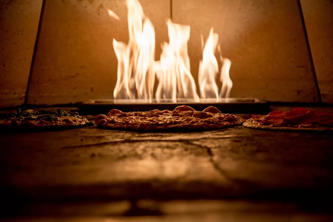Three pizzas bake on the stone floor of a wood-fired oven with bright flames in the background, capturing the essence of luxury lifestyle dining inspired by the vibrant flavors of Majorca.