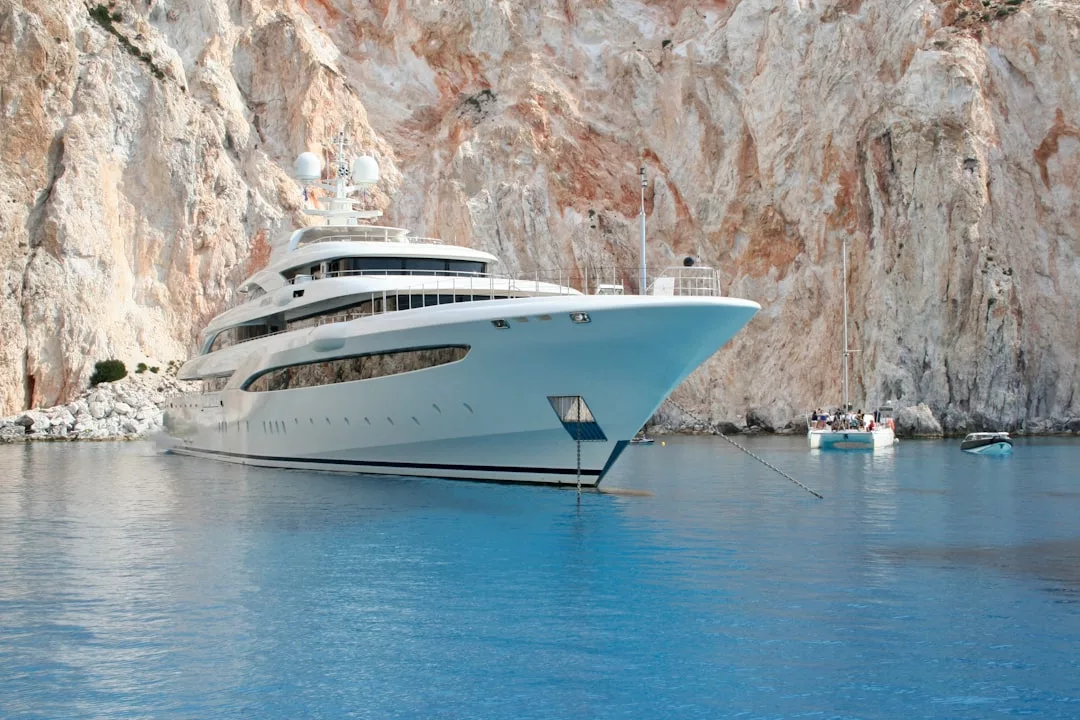 A large white luxury yacht is anchored near a rocky cliff in the calm blue waters of Majorca, with two smaller boats nearby.