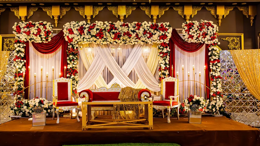 A lavish wedding stage setup with red and gold chairs, a luxury red and gold sofa, floral arrangements, draped fabric, and decorative lighting for an unforgettable celebration.