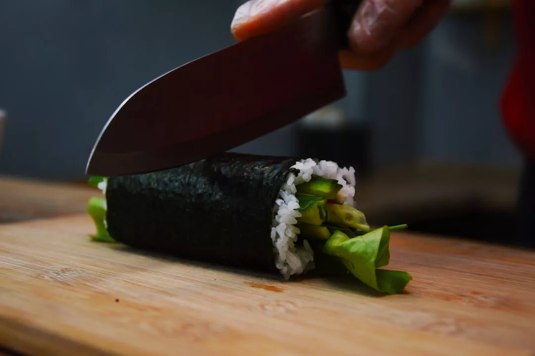A hand with a knife slices a sushi roll filled with rice and green vegetables on a wooden cutting board, capturing a taste of luxury lifestyle inspired by the elegance of Majorca.