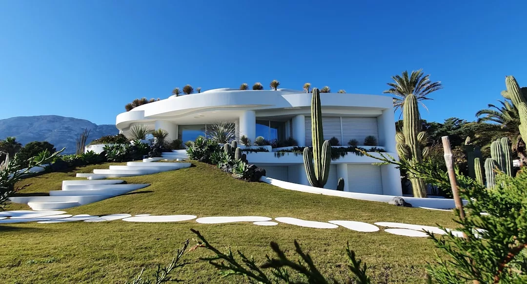 A modern white circular house with large windows offers a taste of luxury living, surrounded by cacti and palm trees on a grassy landscape in Majorca, under a clear blue sky.