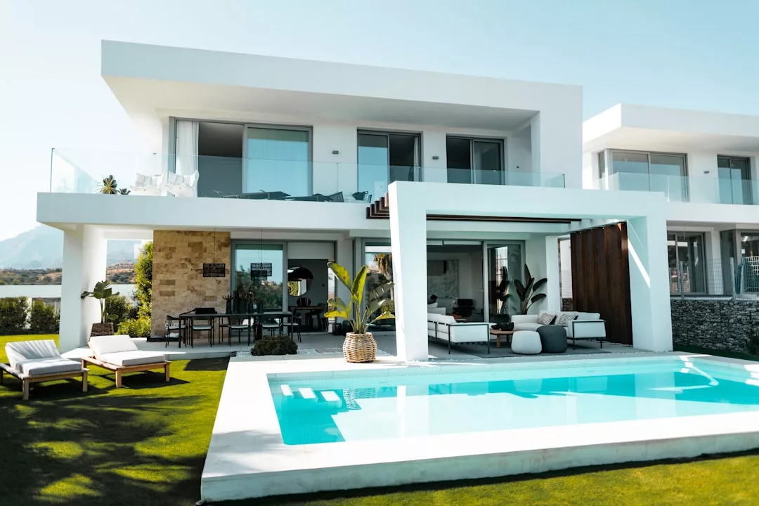 Modern two-story white house with large windows, outdoor patio, pool, and lounge chairs surrounded by a manicured lawn—perfect for enjoying a luxury Majorca lifestyle.