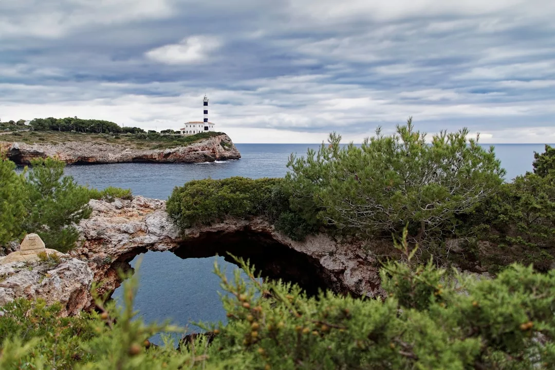 A lighthouse stands on a rocky cliff overlooking the sea in Majorca, with dense green bushes and a natural stone arch in the foreground under a cloudy sky, capturing the island’s unique luxury lifestyle.