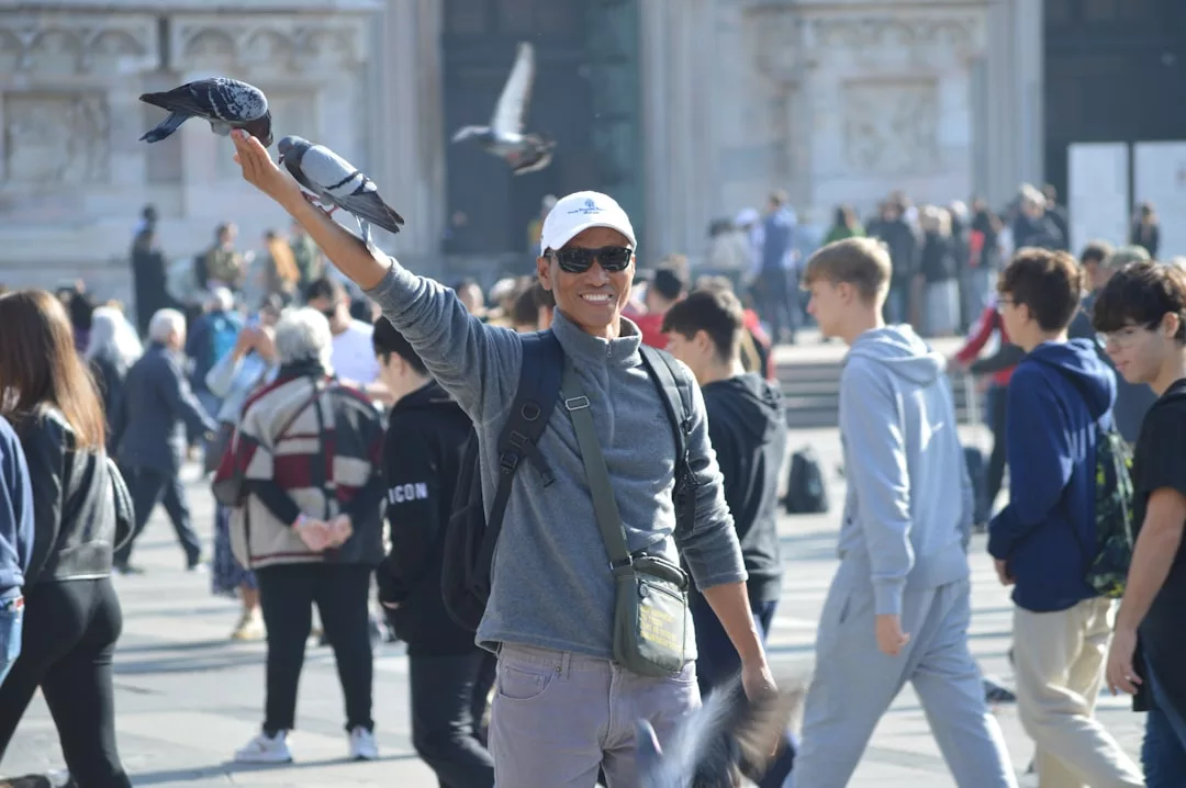 A man wearing sunglasses and a hat stands in a busy square in Majorca, smiling with pigeons perched on his outstretched arm. The lively scene captures the relaxed lifestyle as people walk by in the background.