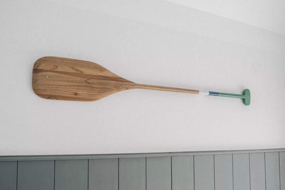 A single wooden canoe paddle with a green and blue handle is mounted horizontally on a white wall above gray paneling, adding a touch of luxury to your lifestyle decor.