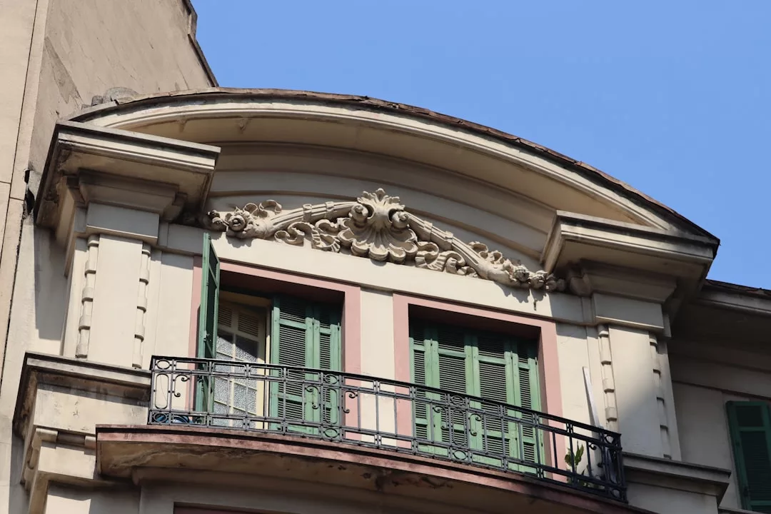 The image shows an ornate building facade in Majorca, featuring a decorative arch, green shutters, and a black wrought iron balcony railing—an elegant expression of Mediterranean luxury against a clear blue sky.