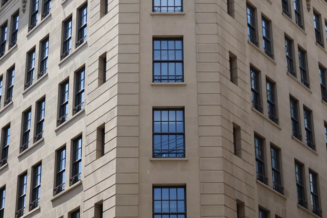 Corner view of a beige stone building in Majorca with tall, rectangular windows reflecting the sky; architectural details include vertical lines and window ledges, capturing a refined sense of luxury and lifestyle.