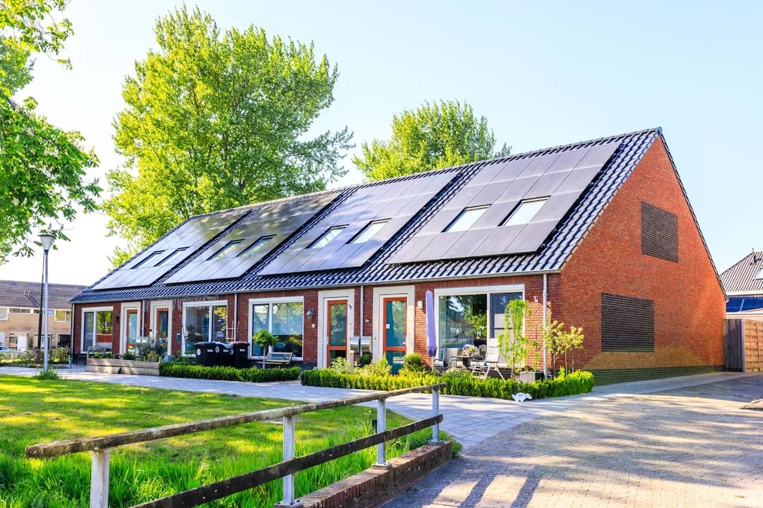 A row of modern brick townhouses with large solar panels on the roofs offers a taste of luxury lifestyle, surrounded by greenery and a paved path under a clear sky.