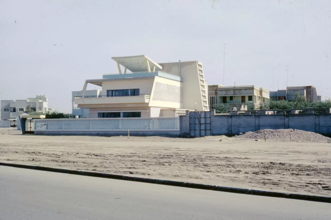 A modern, angular three-story luxury building with a flat roof stands behind a low wall and empty dirt lot, offering an exclusive lifestyle amid other residential buildings in the background.