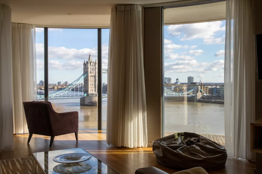 A modern luxury room with large windows overlooks the Thames River and Tower Bridge in London, offering a stylish lifestyle with a chair and beanbag set beside the glass.